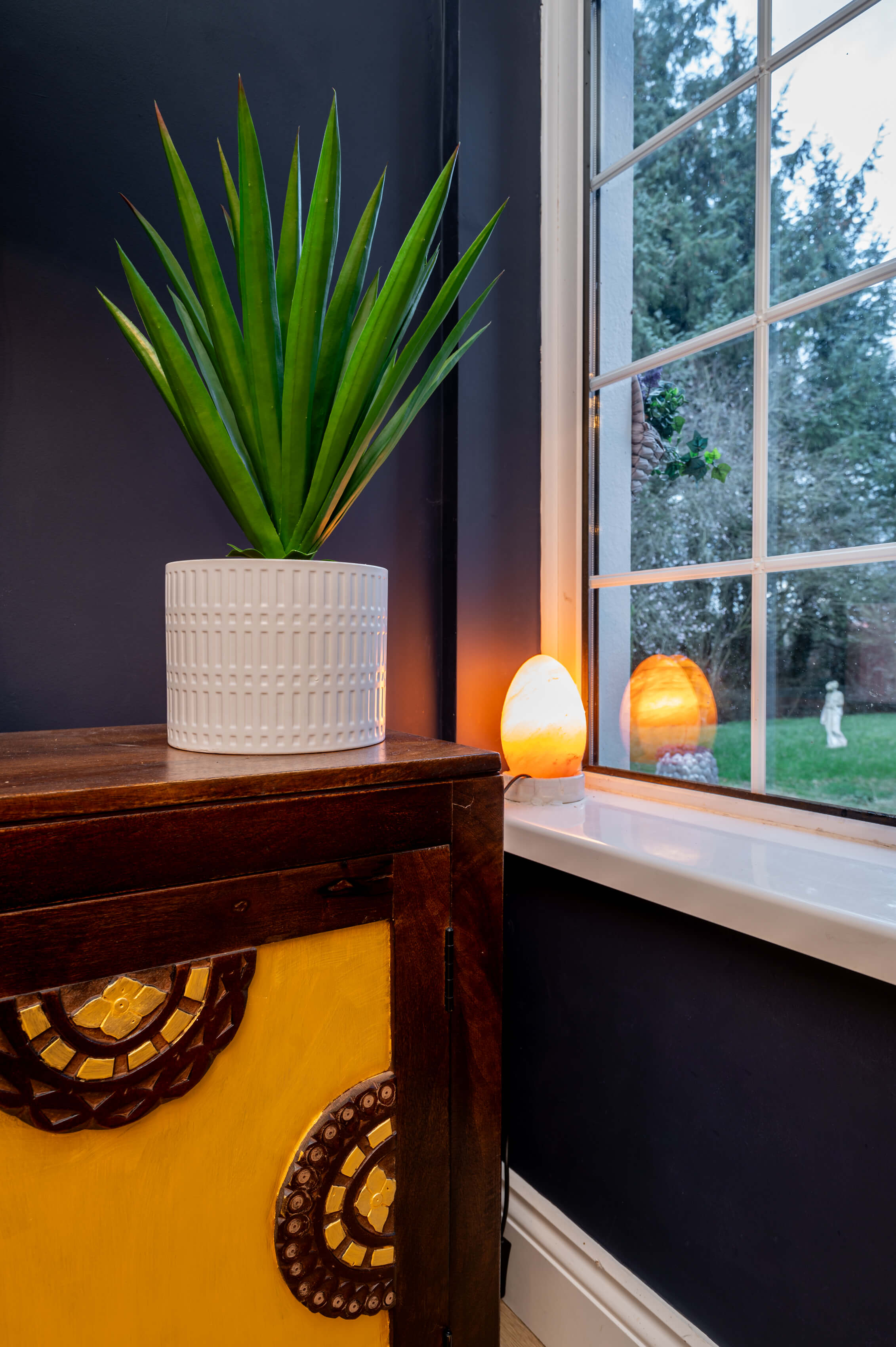 a close up photo of a window and a mango wood storage unit within a living room. There is a white plant pot with greenery and a salt lamp in the window. The unit has been painted with some gold paint.